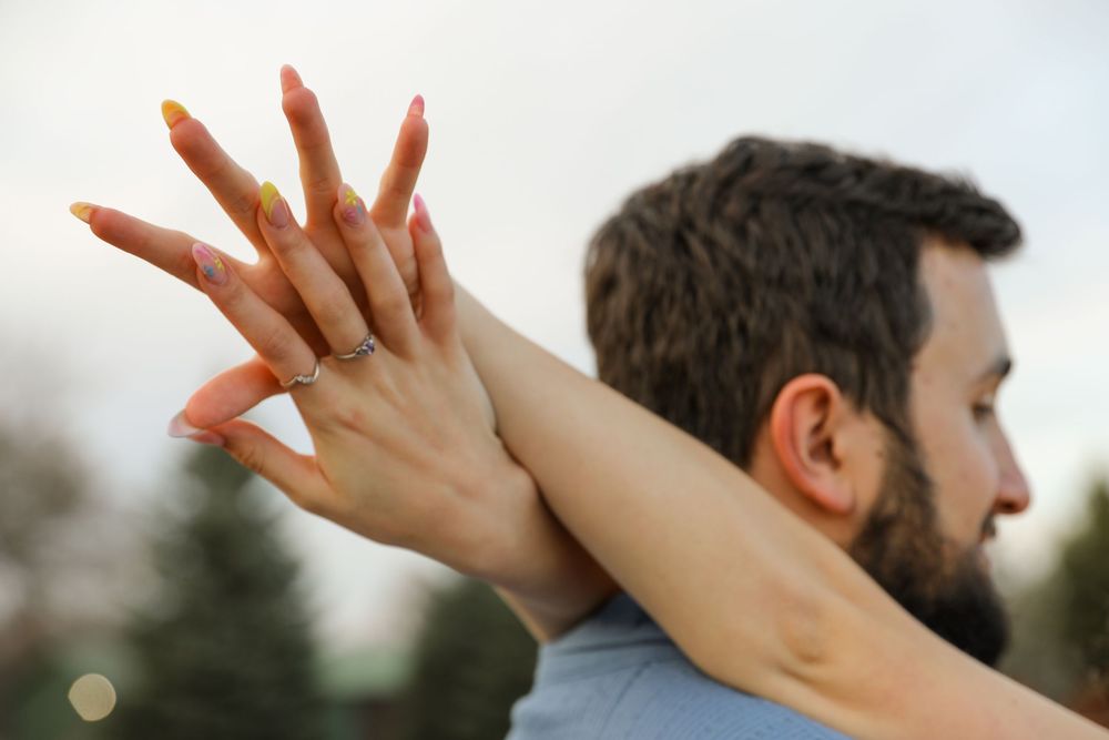 Woman with pink nails and engagement ring embracing a bearded man outdoors in soft natural light.