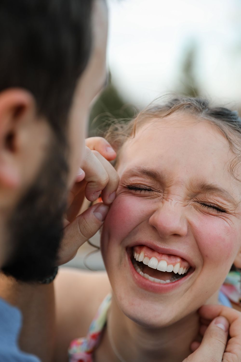 A laughing young woman with curly hair shares a joyful moment with a bearded man outdoors.