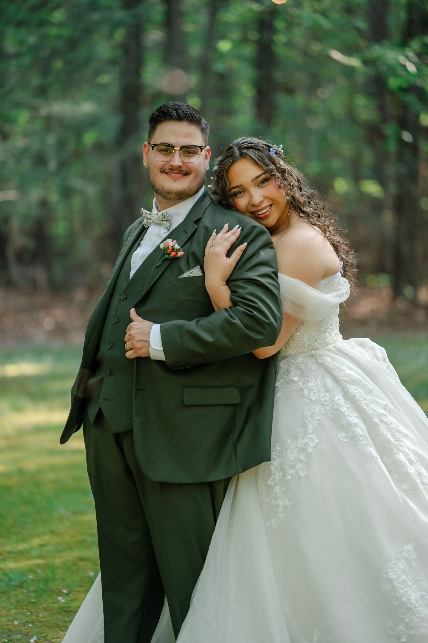 A bride in a white lace ballgown embraces a groom in a dark green suit with bow tie, posing outdoors.