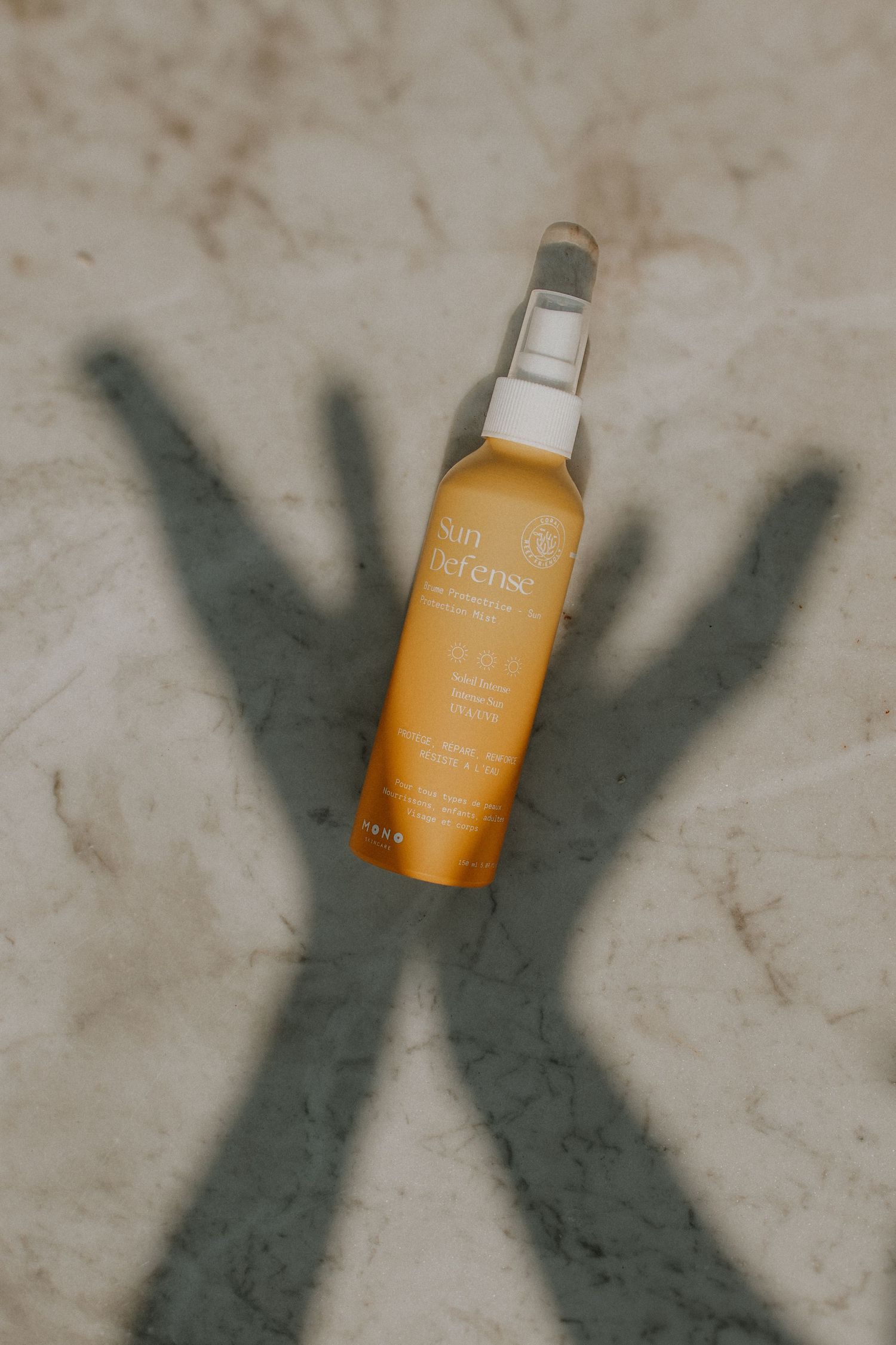 Sun Defense sunscreen spray bottle on marble surface with dramatic shadow of raised hands in background.