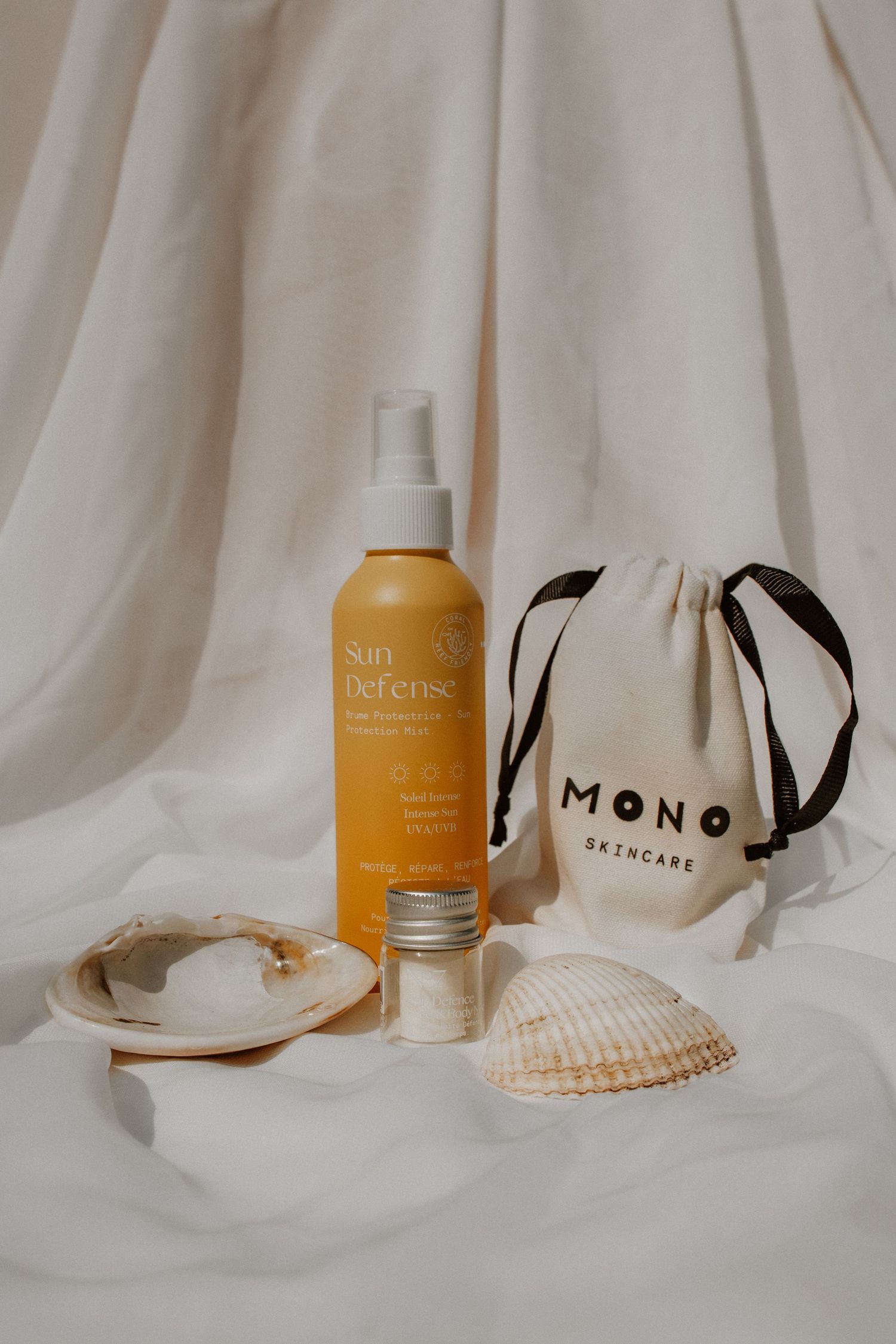 Mono Skincare sun defense spray bottle with seashells and branded cotton drawstring bag on white fabric.