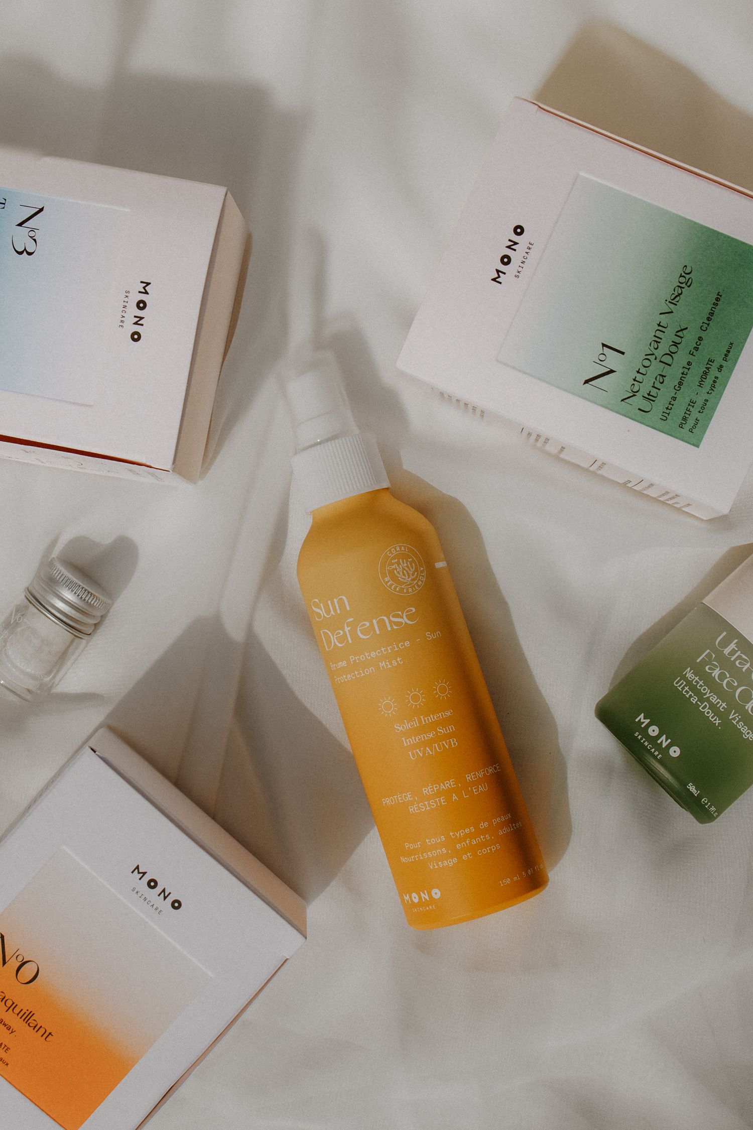 Flatlay of MONO skincare products including Sun Defense spray bottle and boxed items on white fabric.