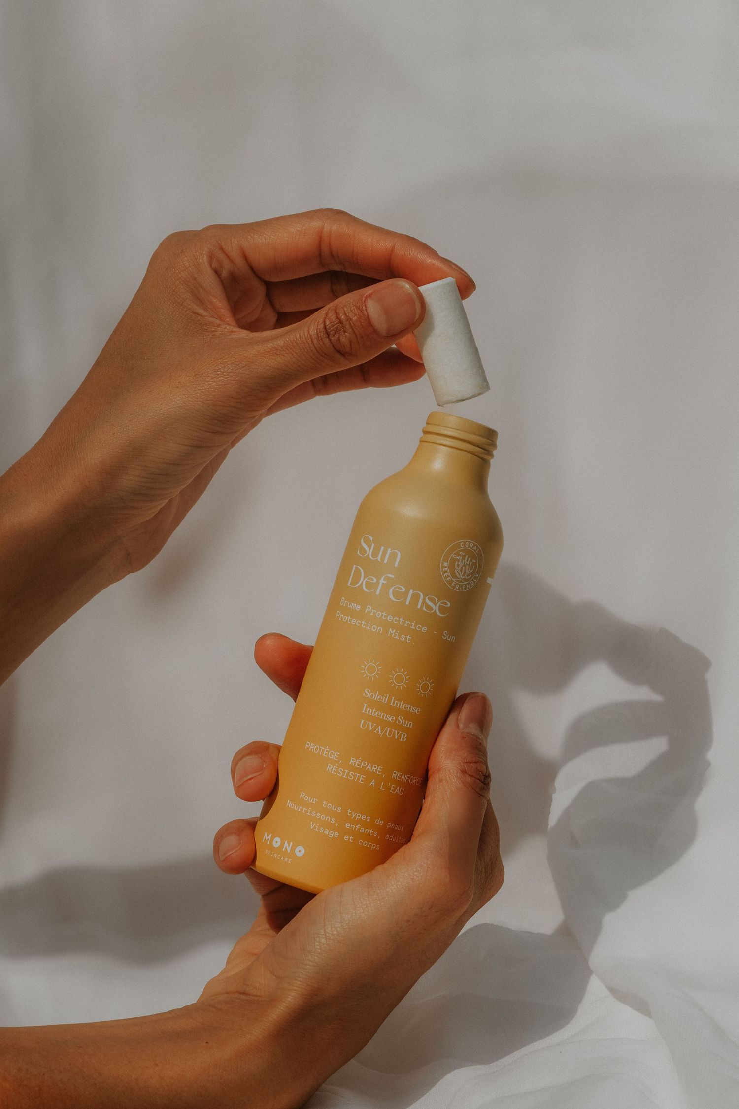 Hands holding a yellow Sun Defense sunscreen bottle with a pump dispenser against a white fabric background.