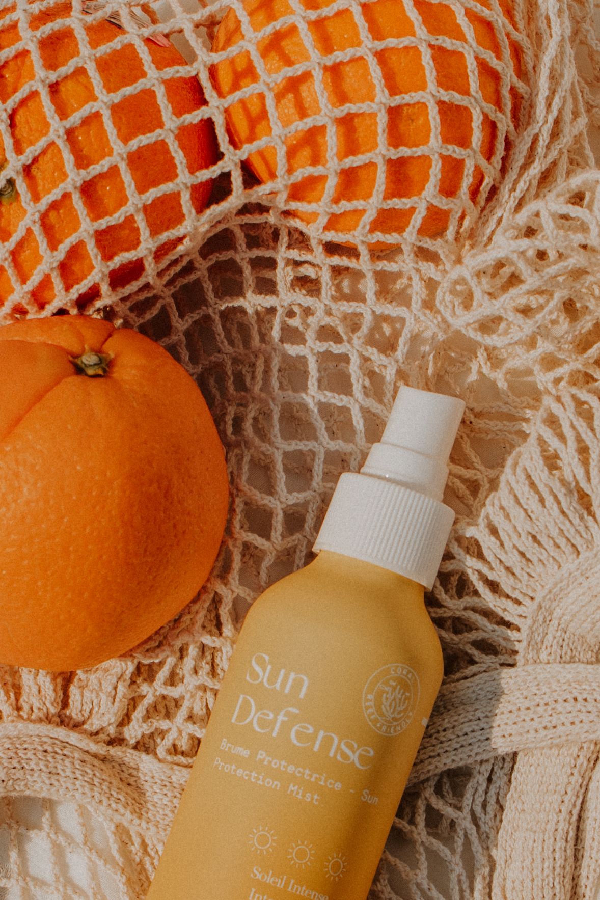 Sun Defense sunscreen mist spray bottle beside fresh oranges in a cream mesh tote bag, summer skincare flatlay.