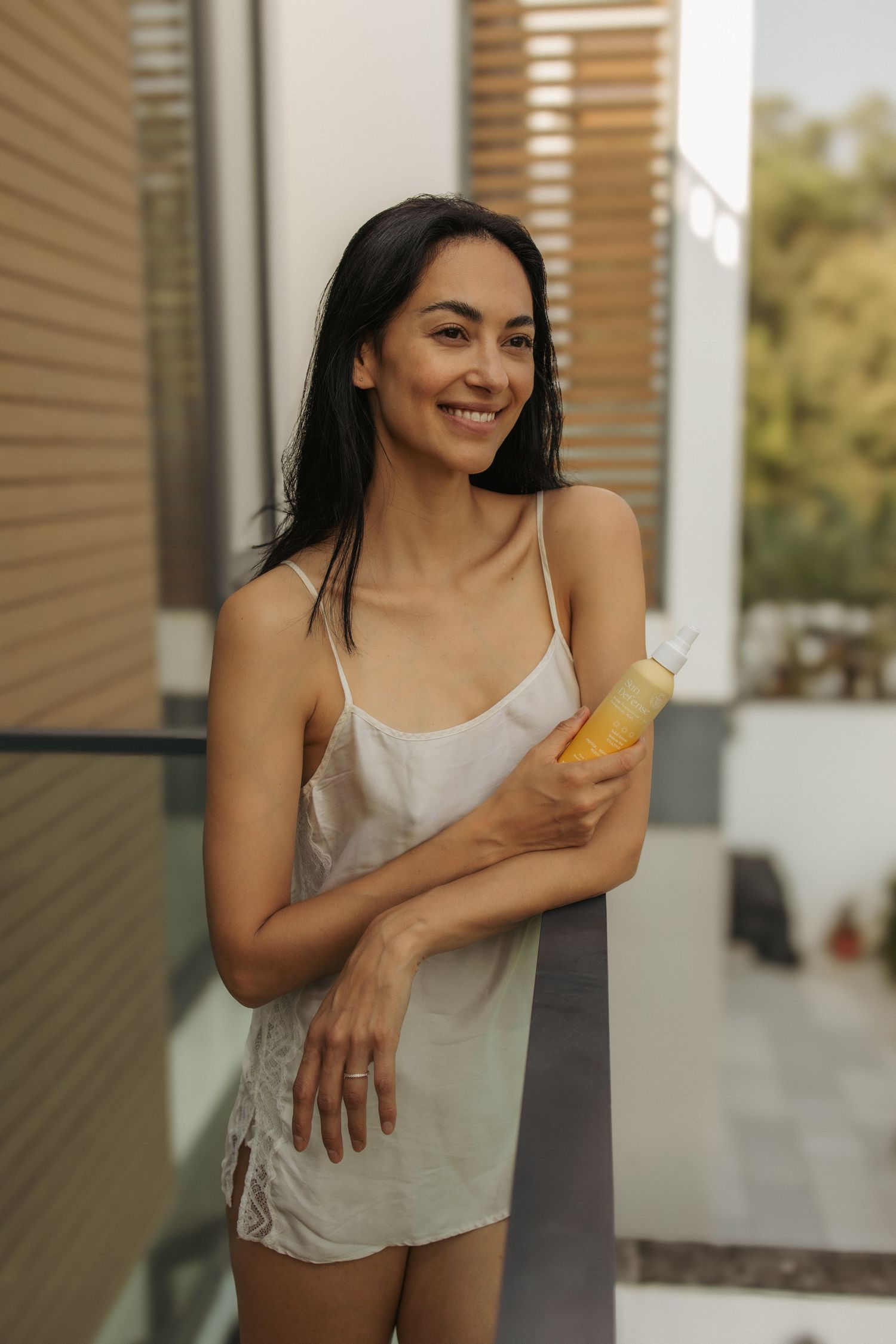 Smiling young woman in white slip dress holding yellow bottle on modern balcony with wooden shutters.