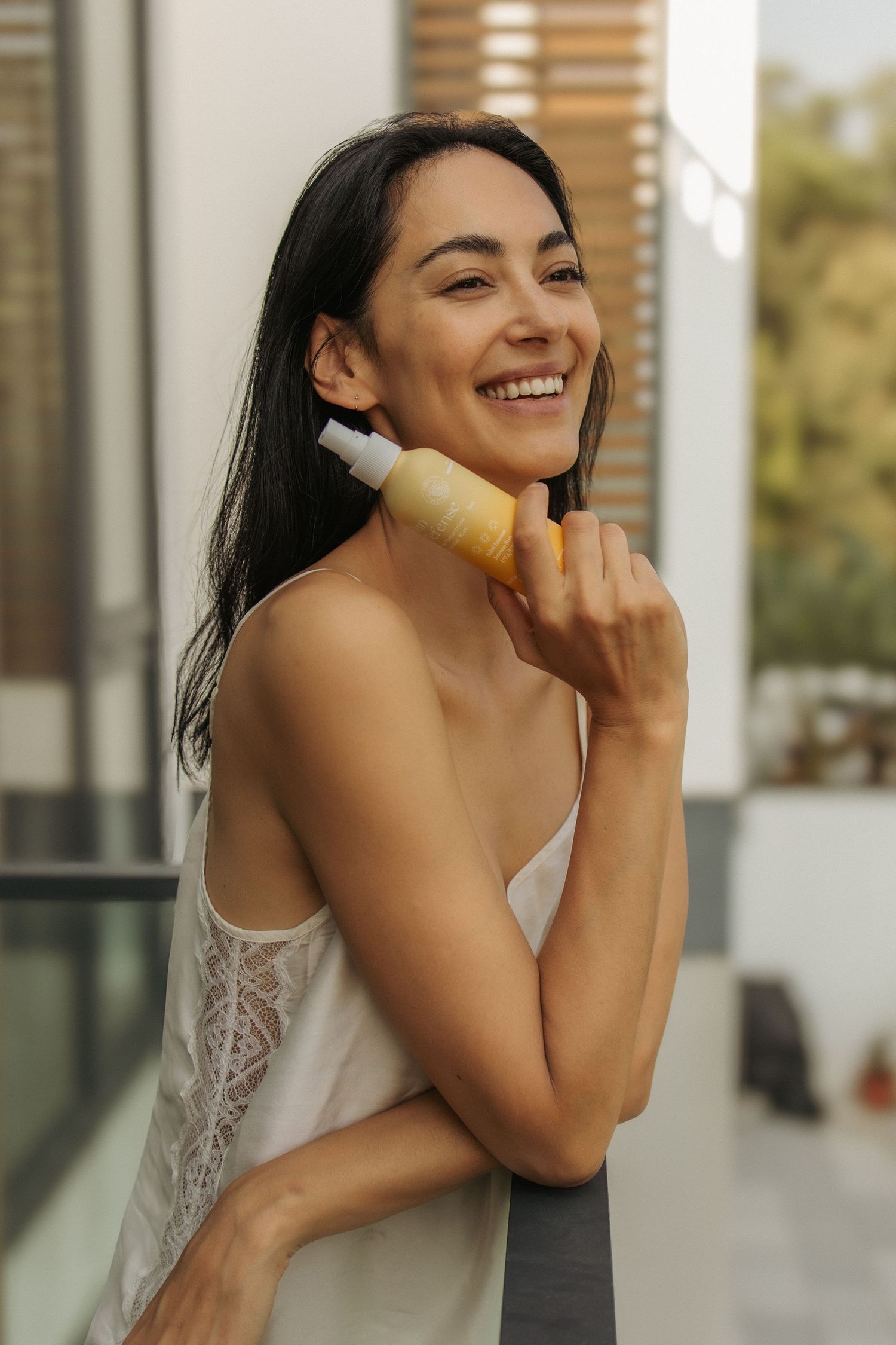 Smiling woman with dark hair applying skincare serum to her face outdoors in a white camisole.