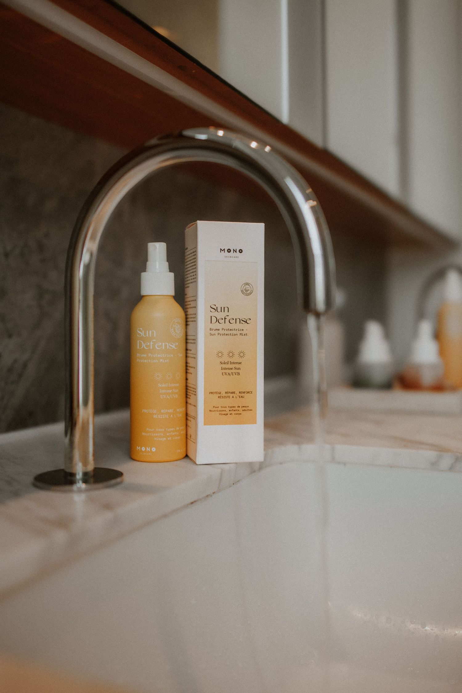 Nene Sun Defense SPF spray bottle and box displayed on marble bathroom countertop near modern chrome faucet.