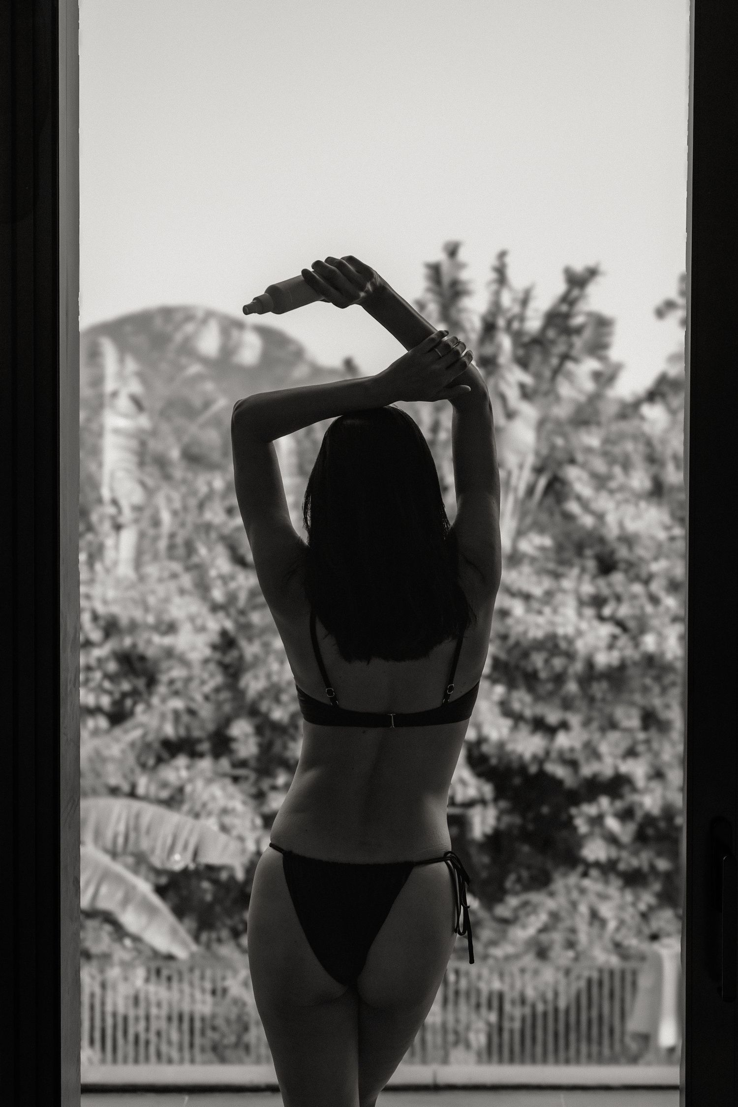 Black and white photo of a woman in a bikini standing by a large window with tropical trees in the background.