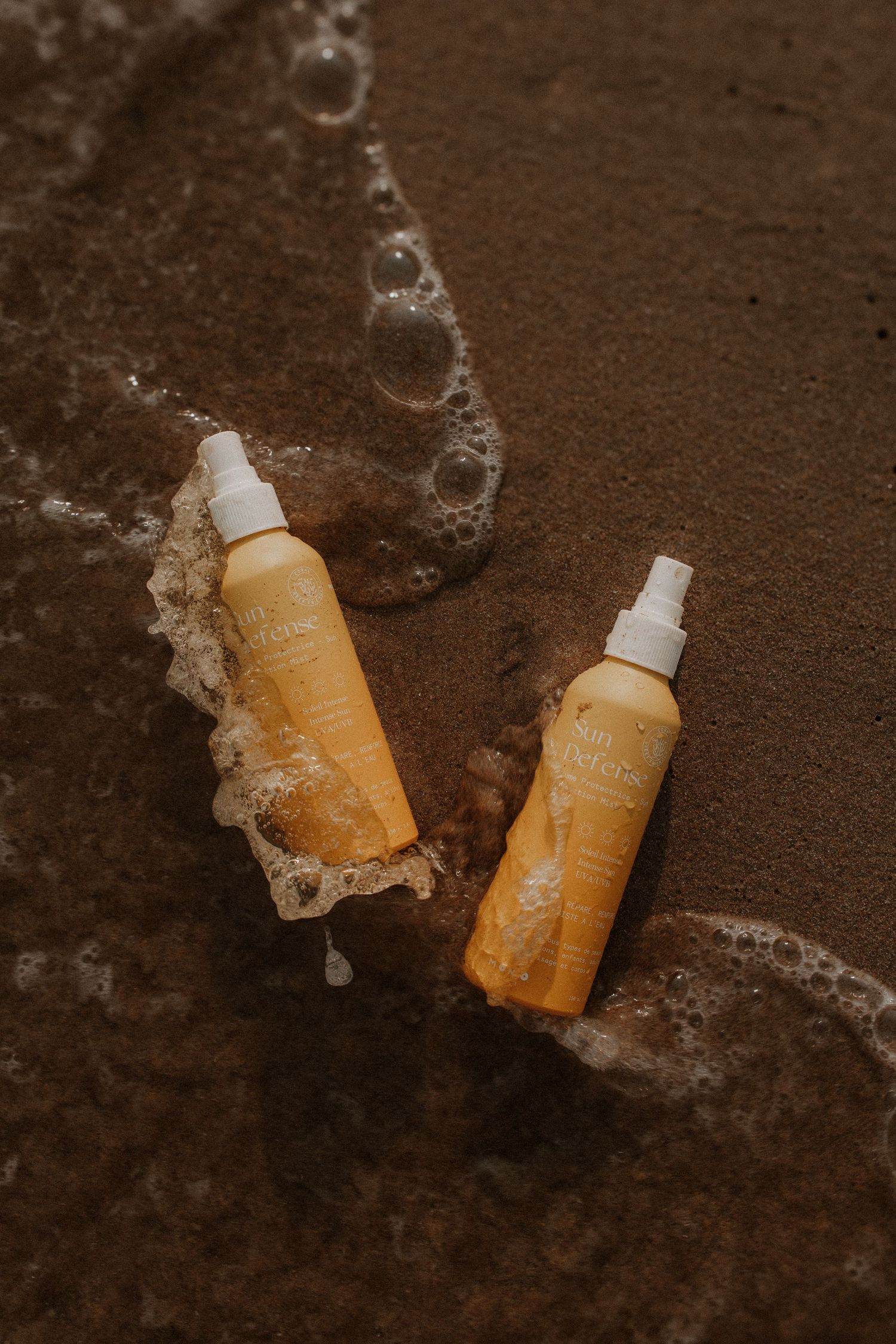 Two yellow Sun Defense spray bottles resting on wet sandy beach with ocean foam waves surrounding them.