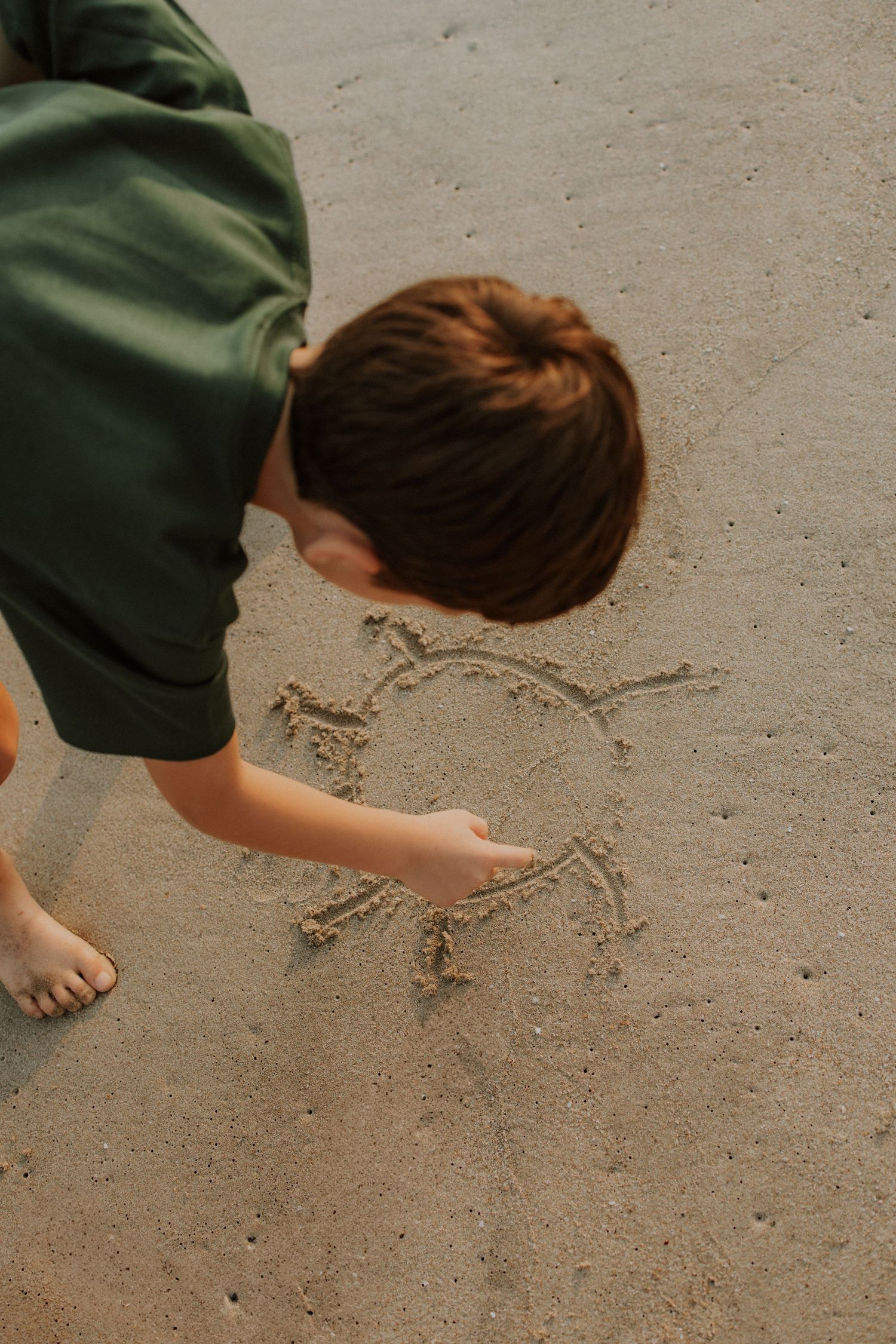 Young boy drawing a sun figure in wet sand at the beach with his finger on a sunny day.