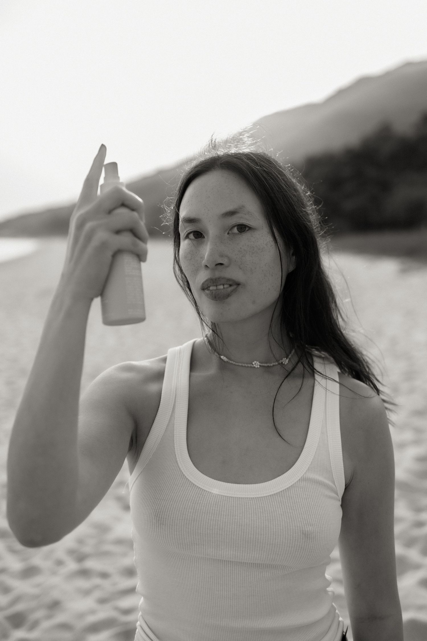 Black and white photo of a woman in a tank top holding a spray bottle on a sandy beach with mountains in background.