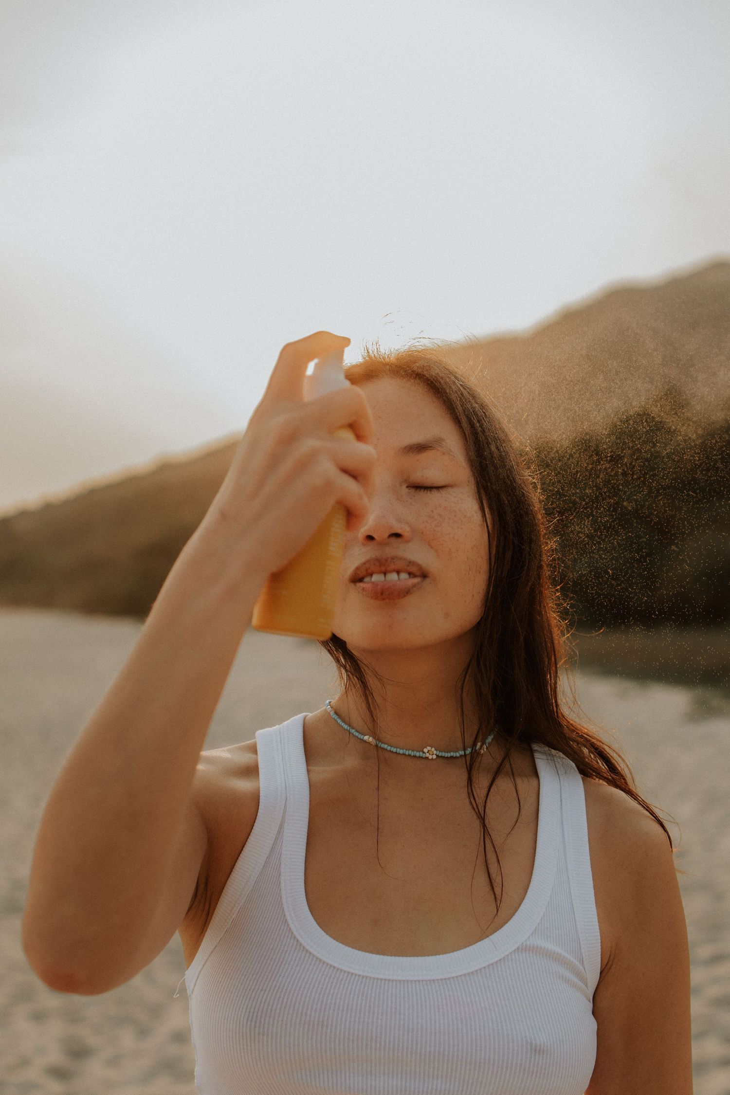 Woman applying sunscreen spray at beach, eyes closed, wearing white tank top with choker necklace, sandy dunes background.