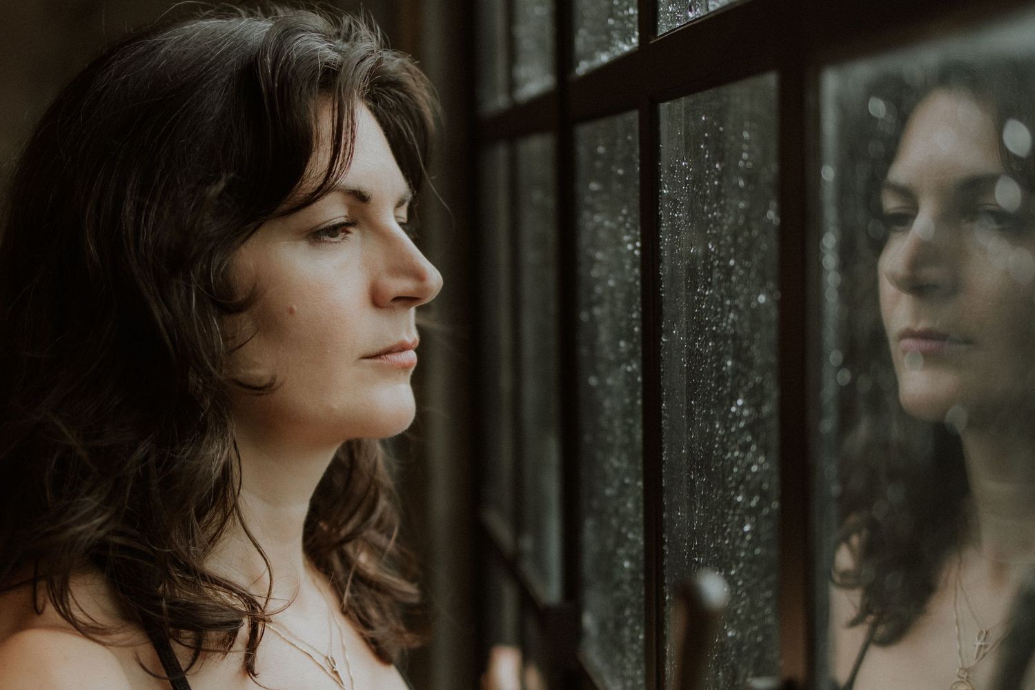 A contemplative woman gazes out a rain-speckled window, her reflection visible in the glass.