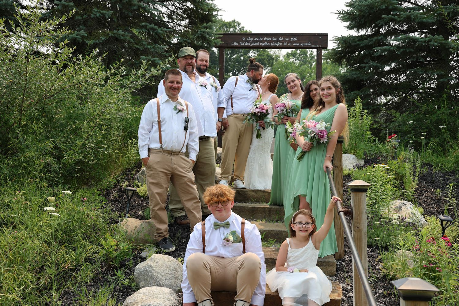 Wedding party poses on garden steps in mint green and khaki attire, surrounded by lush greenery.