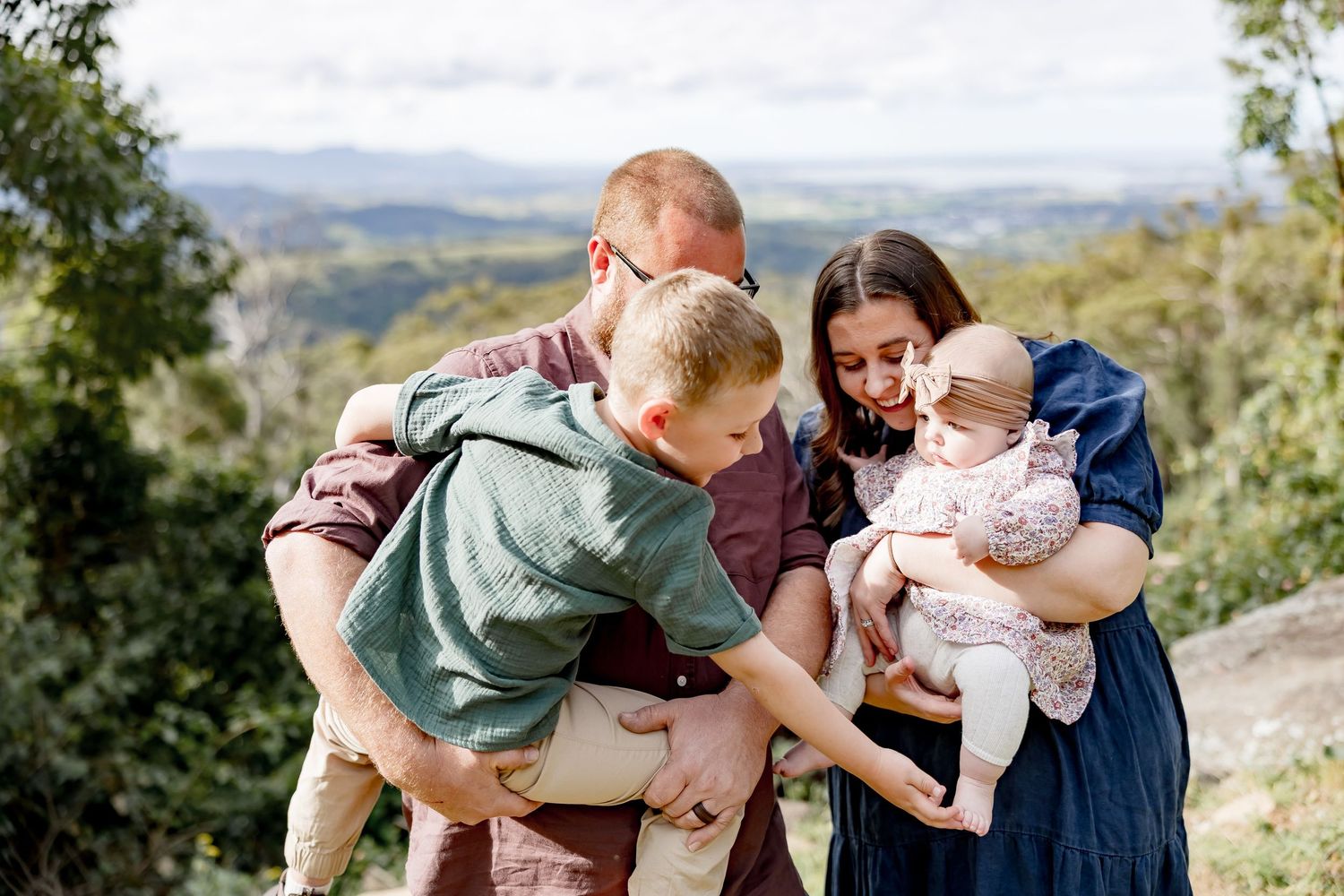 Family of four outdoors with scenic valley views, parents holding baby girl while young boy leans in for a closer look.