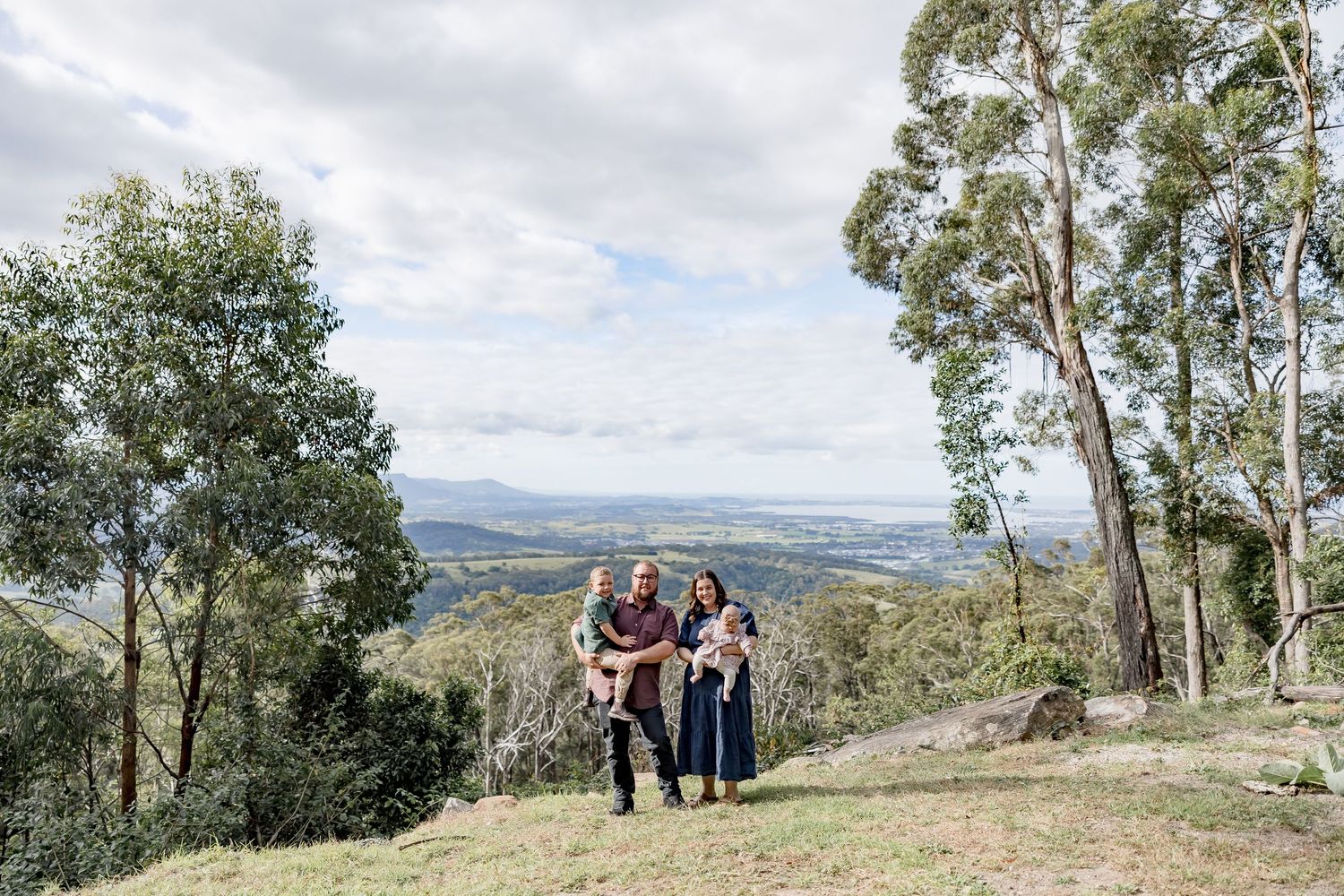 Family of four posing on a scenic hilltop surrounded by eucalyptus trees with panoramic valley views.