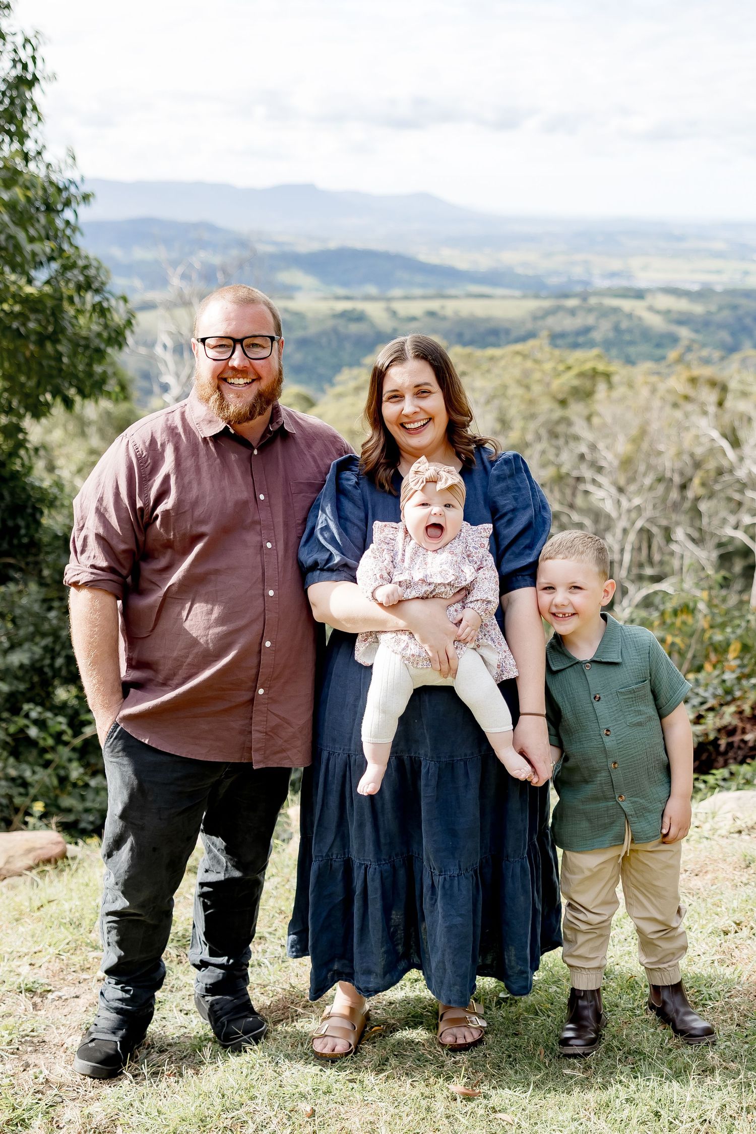 Happy family of four posing outdoors with scenic mountain valley views, parents holding young children on a hilltop.