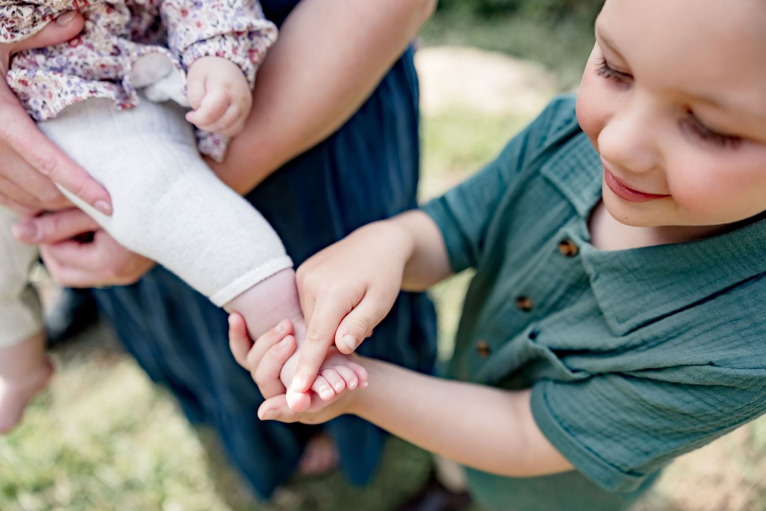 Young boy in green shirt gently holds baby sibling's hand outdoors, captured in a tender family moment.