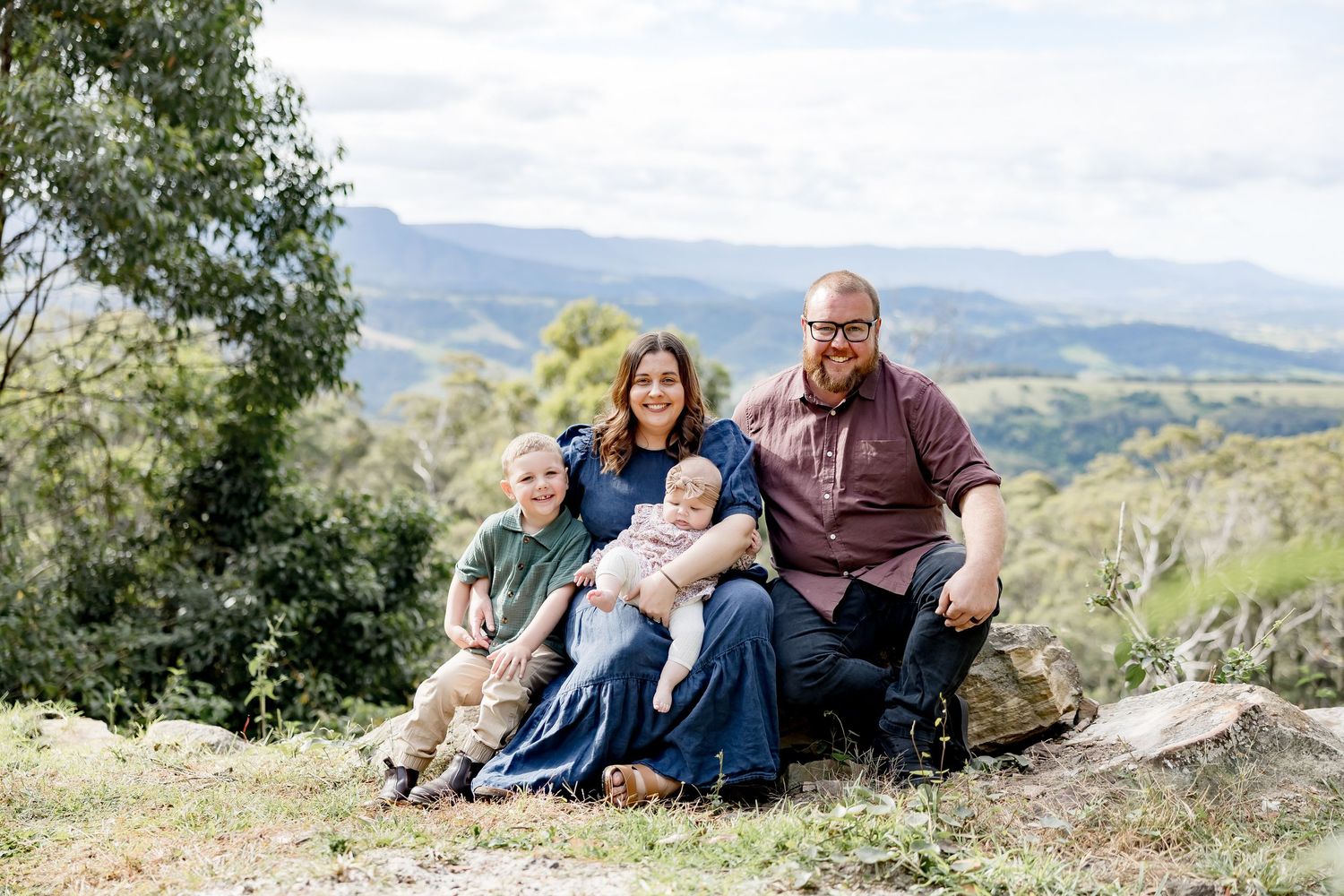 Family of four sitting on rocks with scenic mountain valley views in an outdoor portrait session.