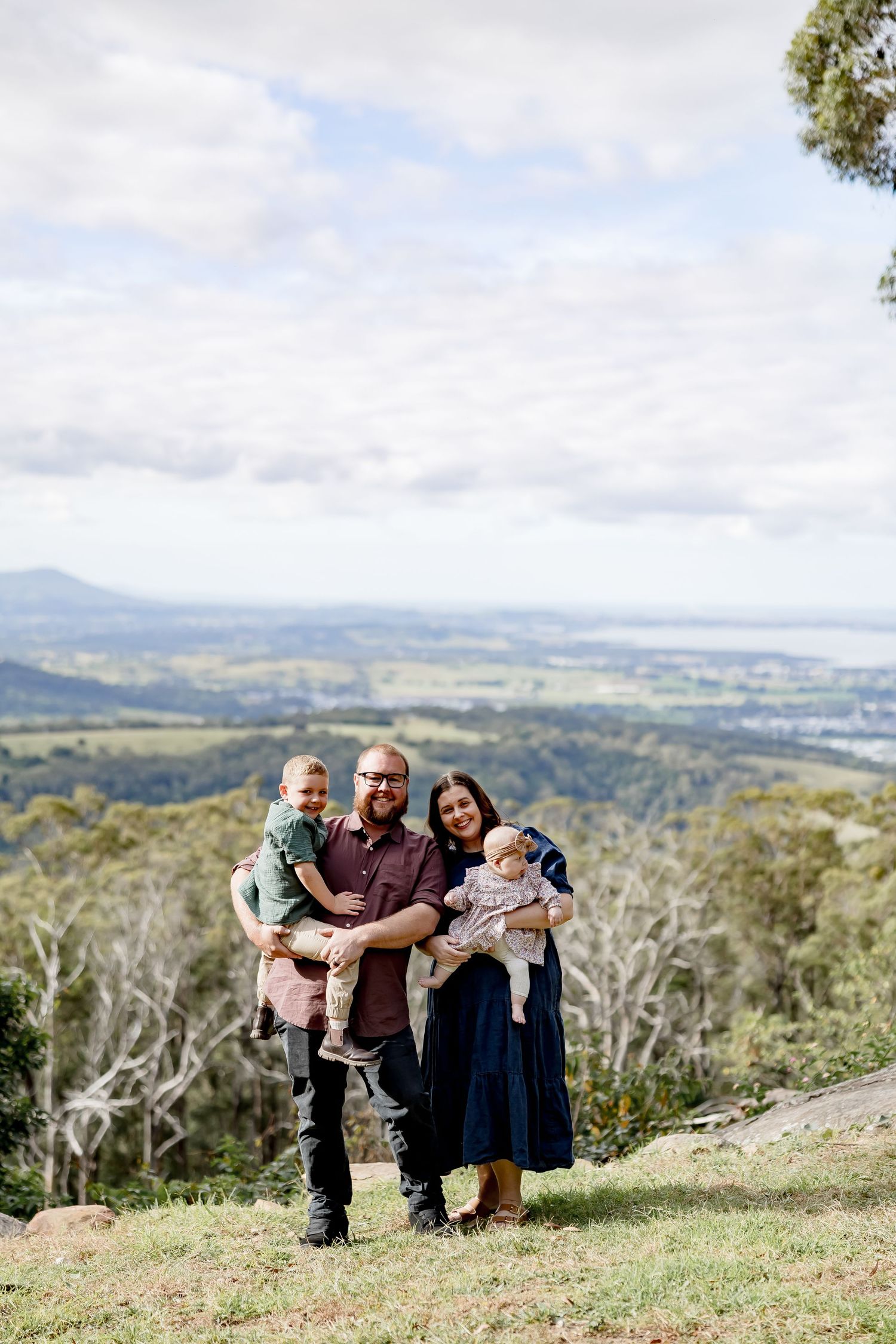 Family of four posing on a hilltop with scenic valley and coastline views on a partly cloudy day.