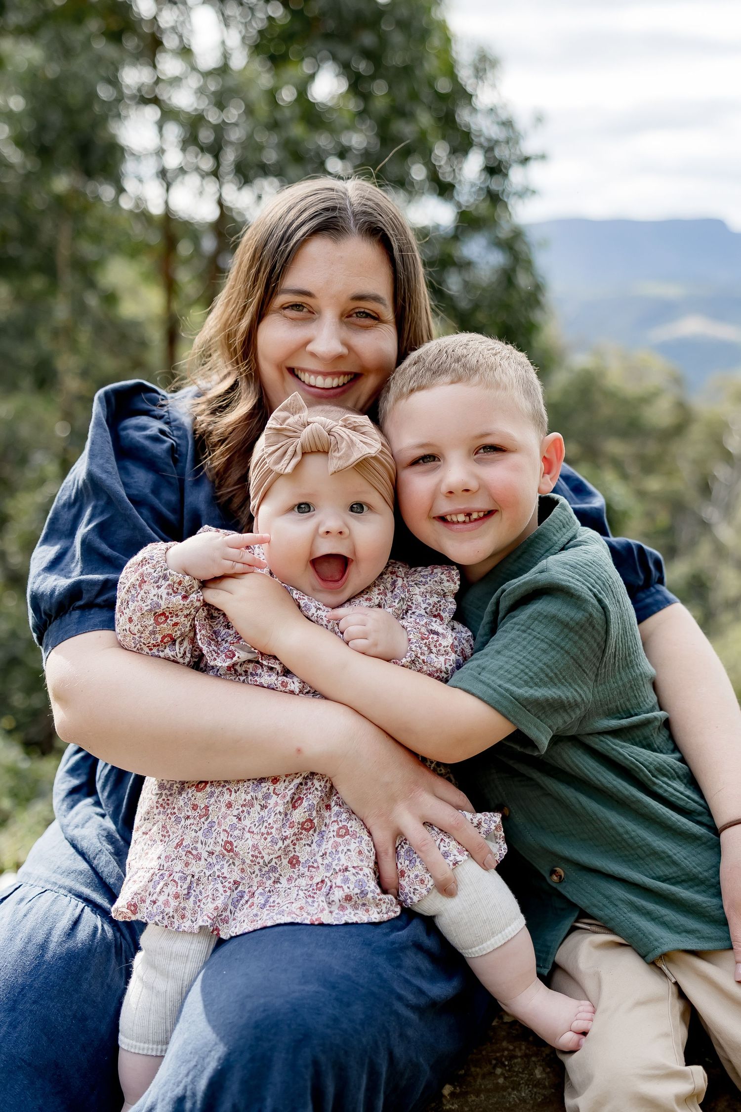 A smiling mother hugs her baby girl and young boy outdoors with mountain views behind them.