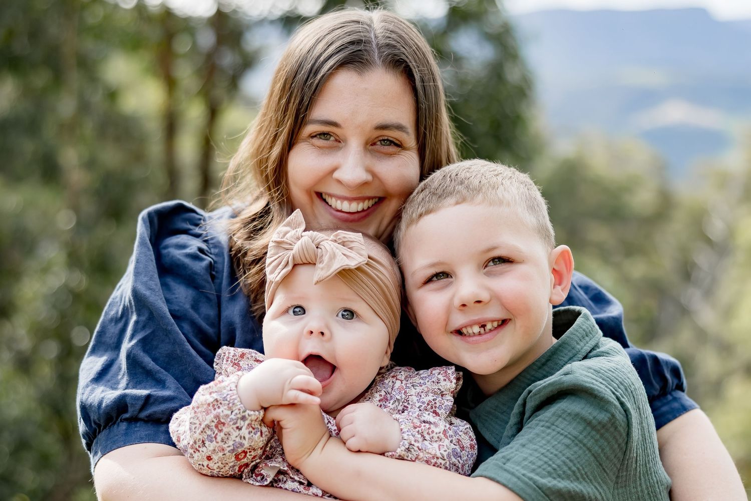 Smiling mother hugging her baby girl and young boy outdoors with green trees and mountains in background.