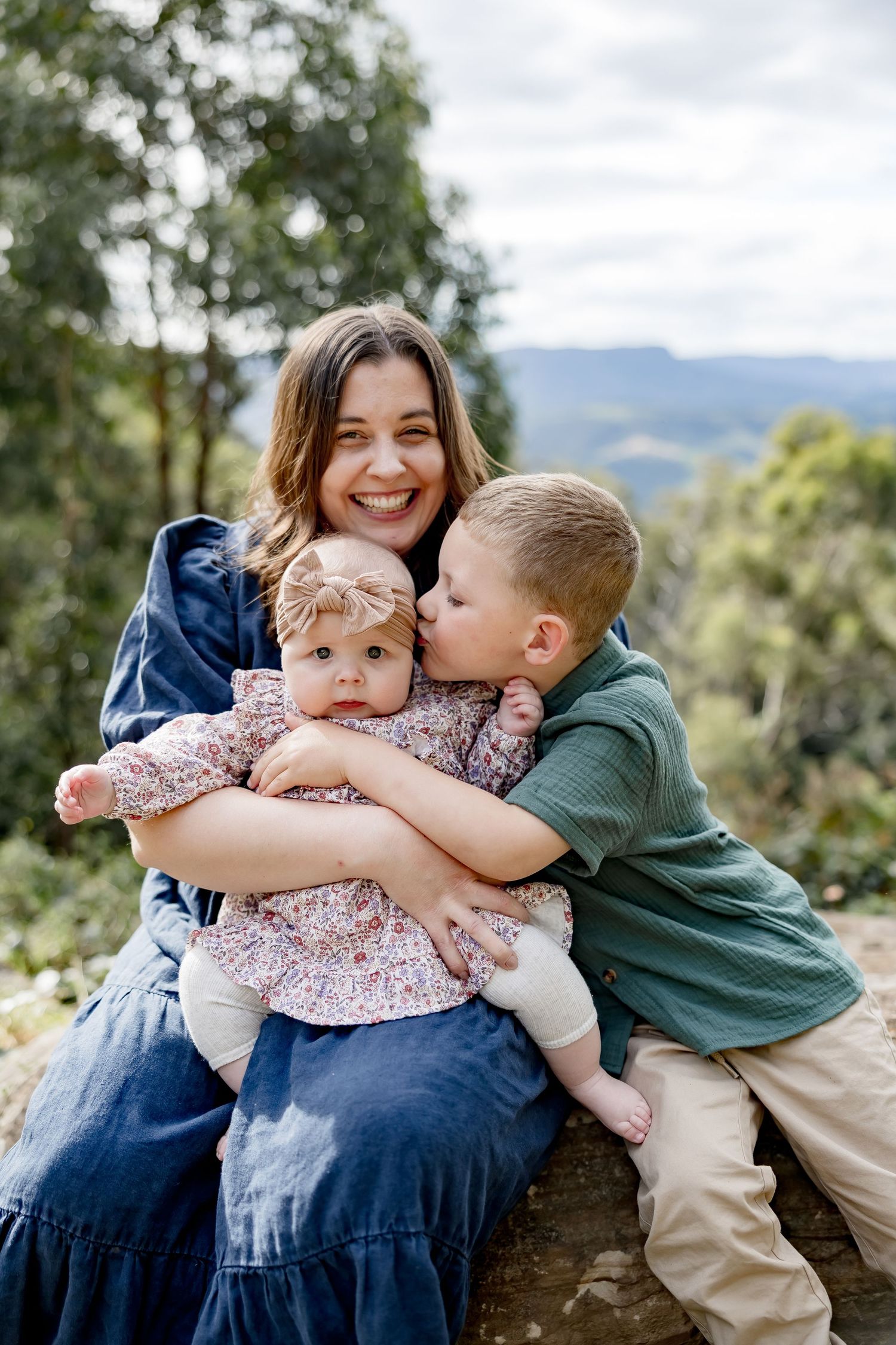 Smiling mother holds baby girl while young boy kisses her cheek outdoors with mountain views.