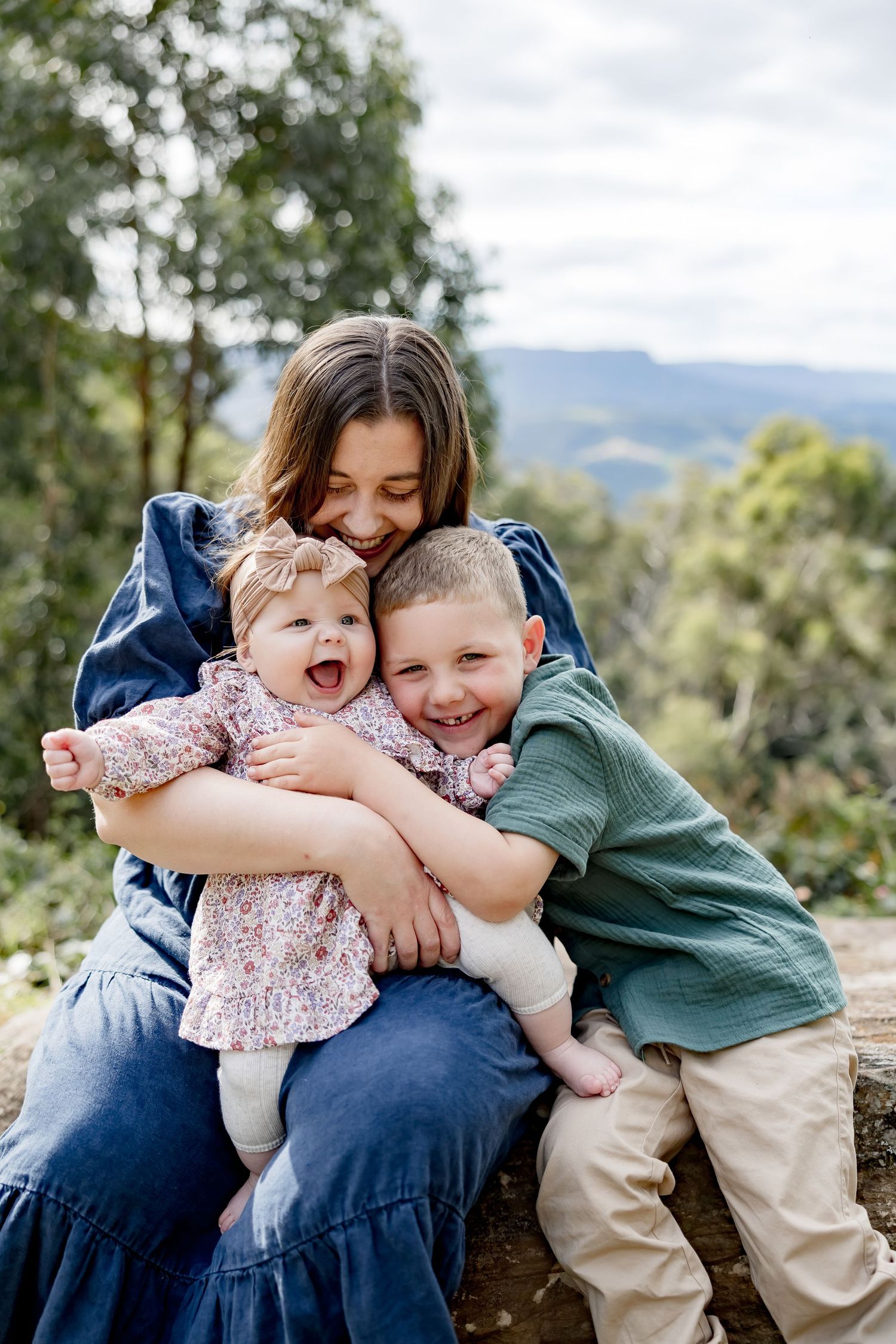 Mother laughing while holding baby girl and hugging young boy outdoors with mountain views.