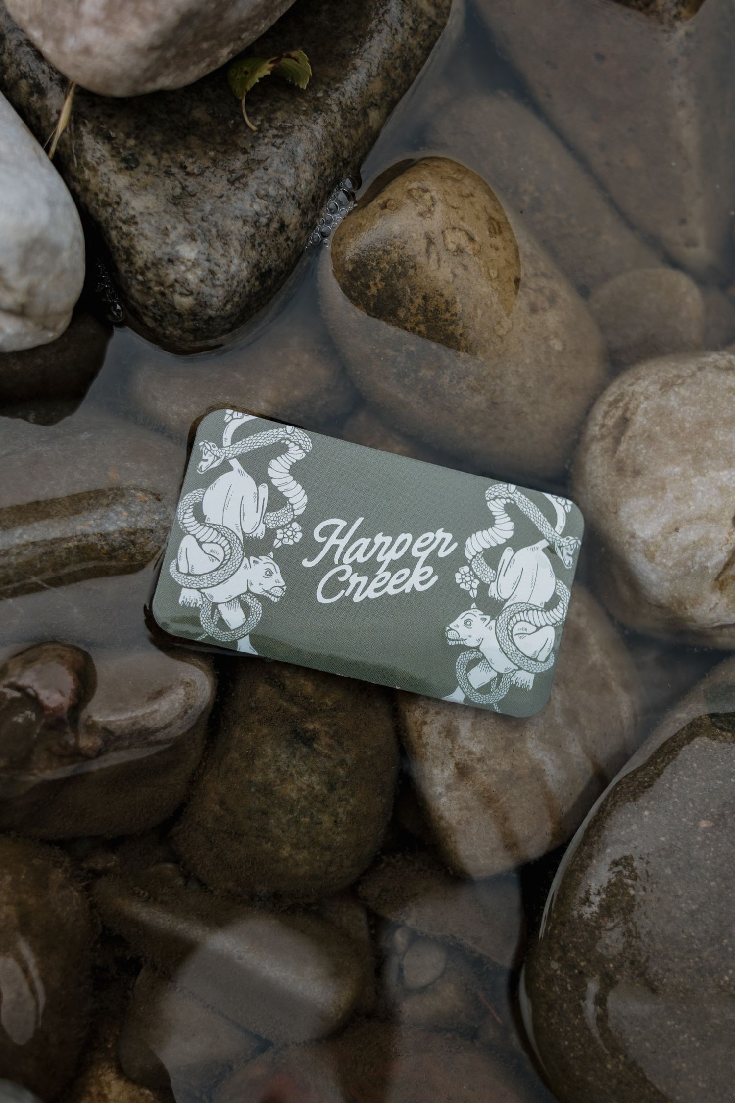 Harper Creek branded soap bar resting on smooth river rocks in shallow water, featuring elegant turtle illustrations.