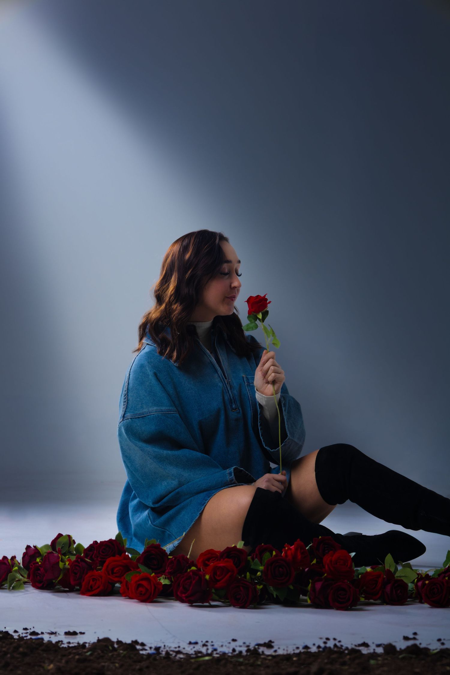 Young woman in blue jacket smelling a red rose, sitting among scattered roses on a studio floor.