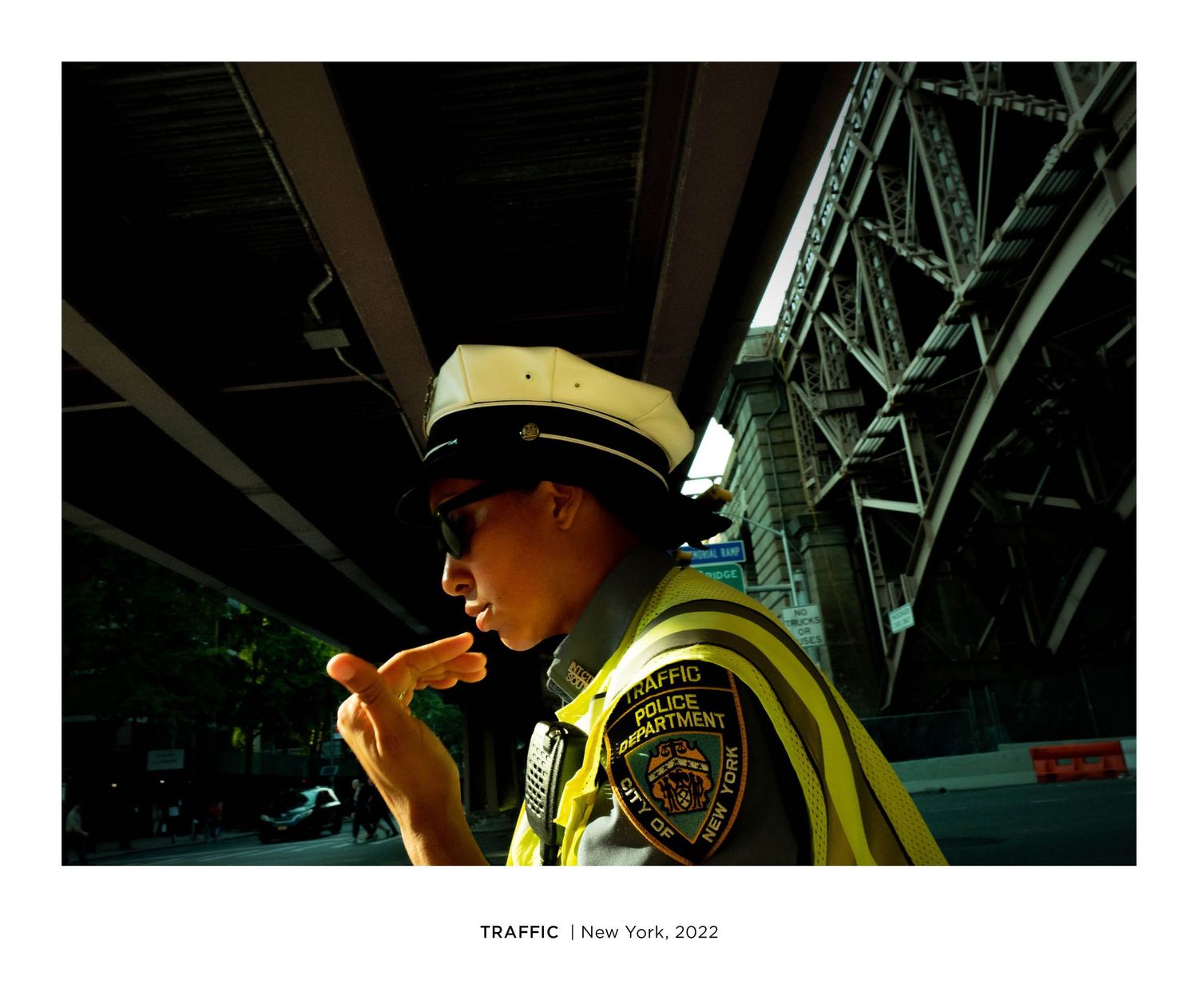 A NYC traffic officer in a yellow vest and white cap stands under a bridge, touching her face, New York, 2022.