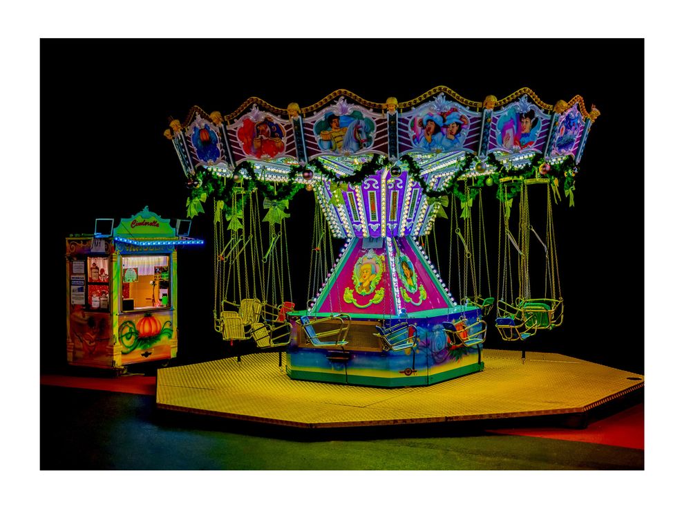 Colorful illuminated carnival swing ride glowing at night with empty seats and a ticket booth beside it.