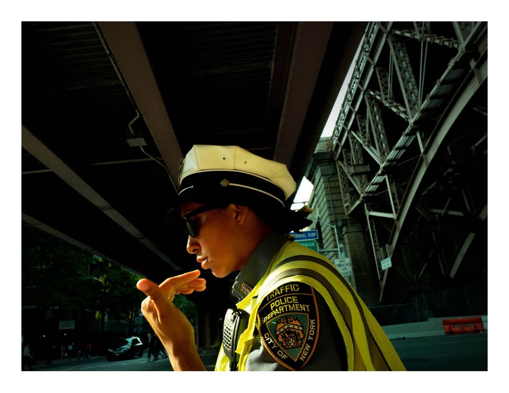 Color photo of a female traffic officer in uniform and reflective vest eating under an elevated structure.