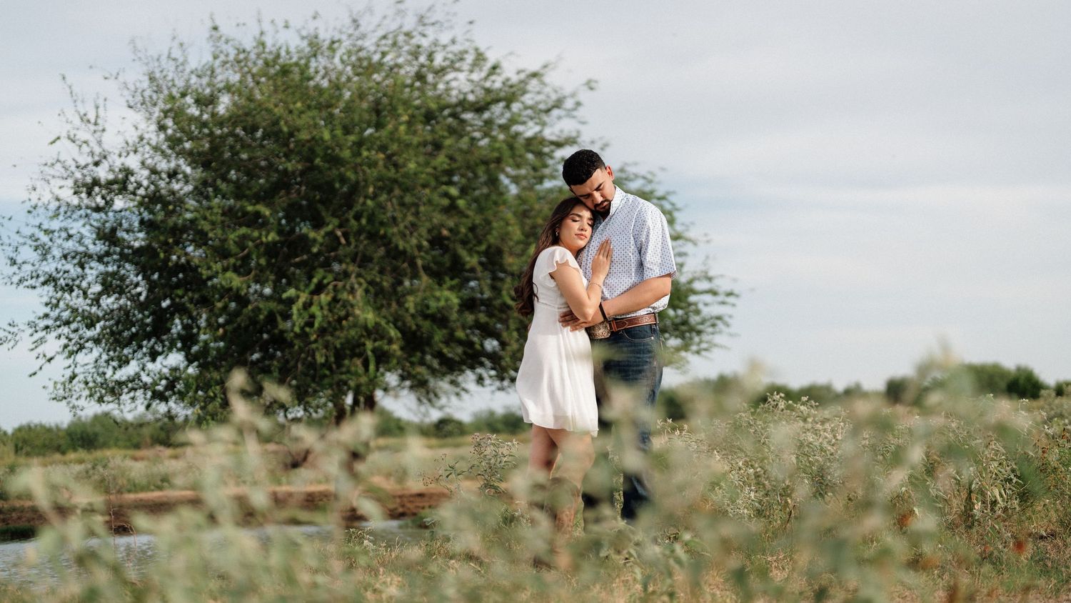 A couple embraces romantically in a lush green field, with the man holding the woman close near a large tree.