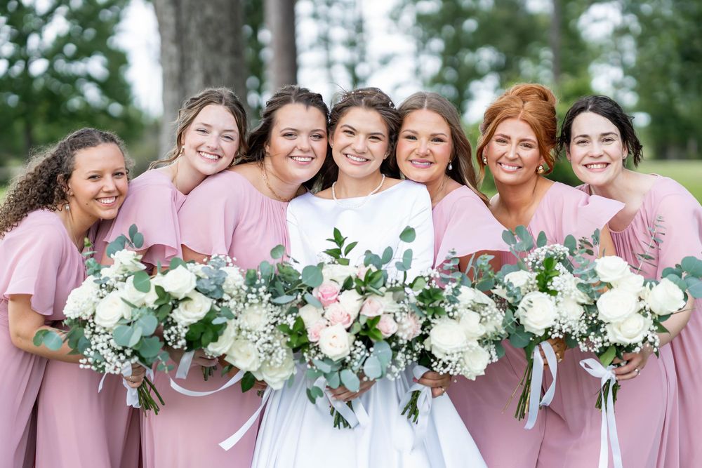 Bride in white gown poses with six bridesmaids in pink dresses holding white and green bouquets outdoors.