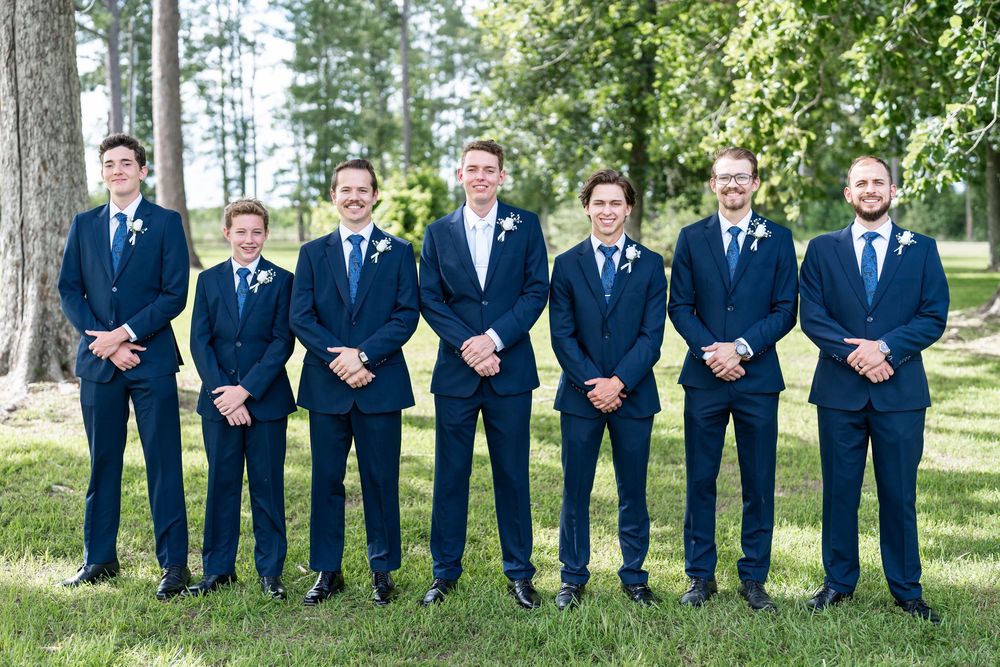 Seven groomsmen in matching navy blue suits posing outdoors on green grass with trees in the background at a wedding.