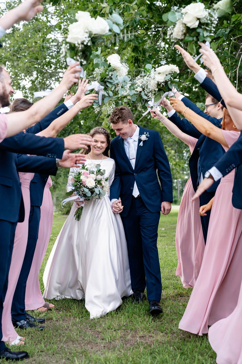 Bride and groom walk through floral arch created by bridesmaids in pink dresses and groomsmen in navy suits outdoors.