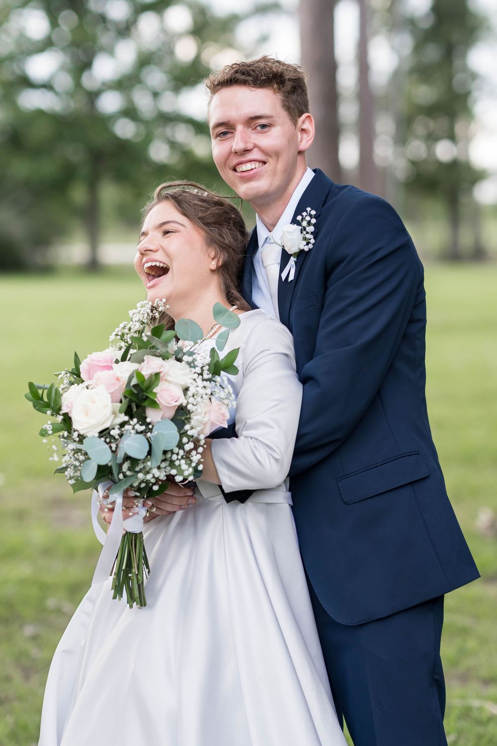 Joyful bride holding pink and white bouquet laughs as groom in navy suit embraces her outdoors.