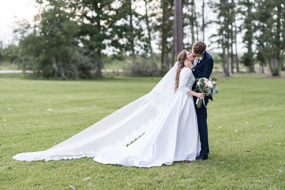 Bride and groom share a romantic kiss on green lawn, her long white veil flowing in the breeze, with tall pine trees behind.