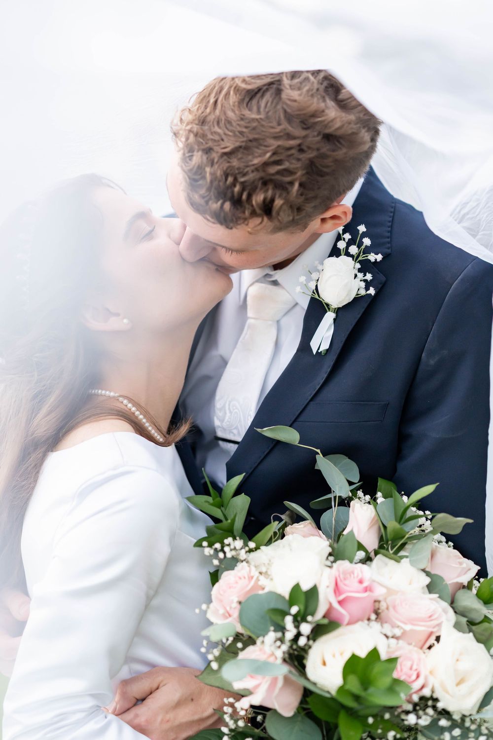 Bride and groom sharing a romantic kiss under a wedding veil, holding a bouquet of pink and white roses.