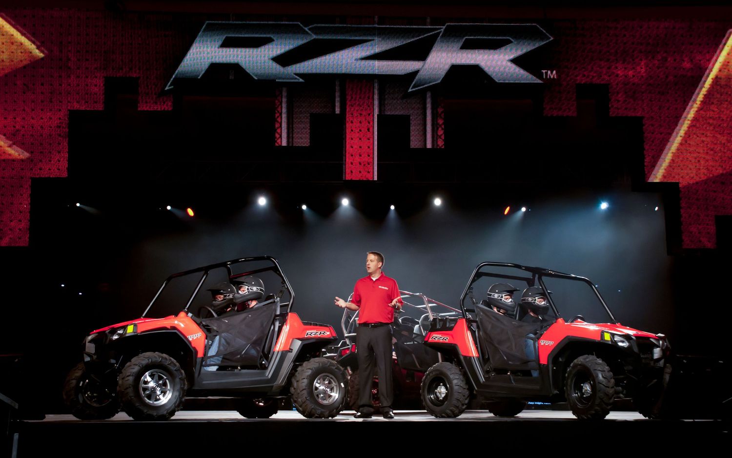 Man presenting two red Polaris RZR side-by-side UTVs on a stage with dramatic RZR logo backdrop.