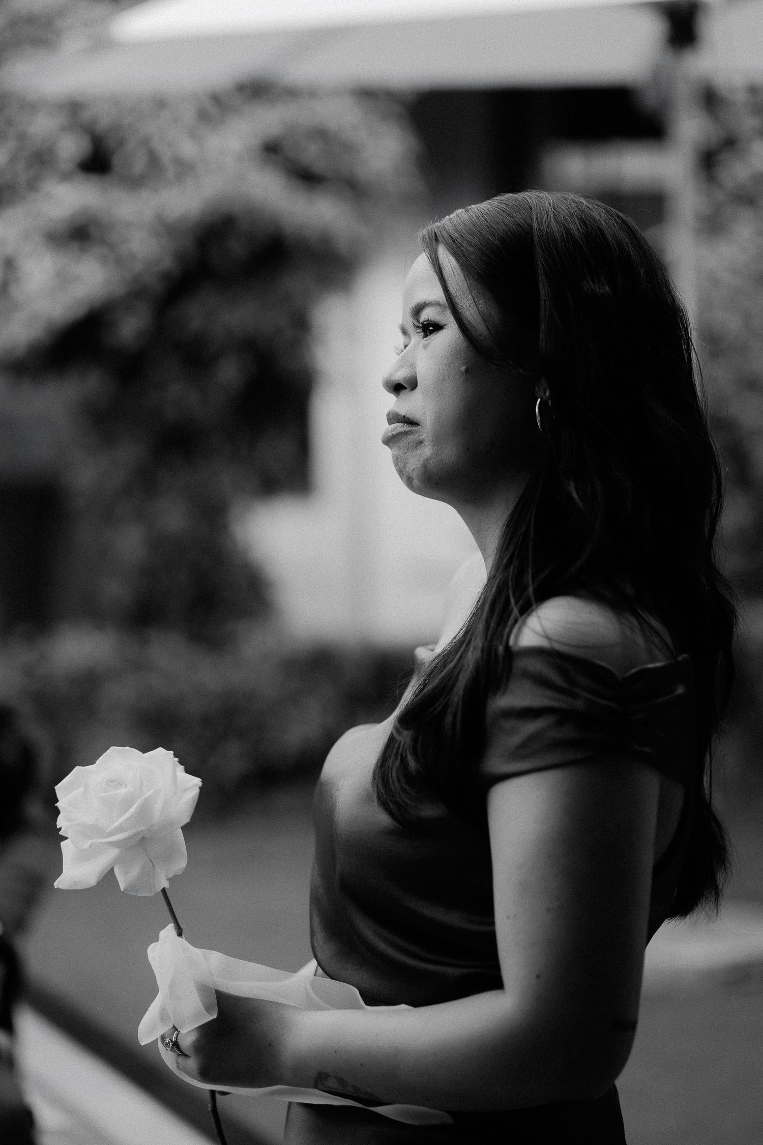 Black and white portrait of a young woman holding a white rose, gazing thoughtfully in an outdoor setting.