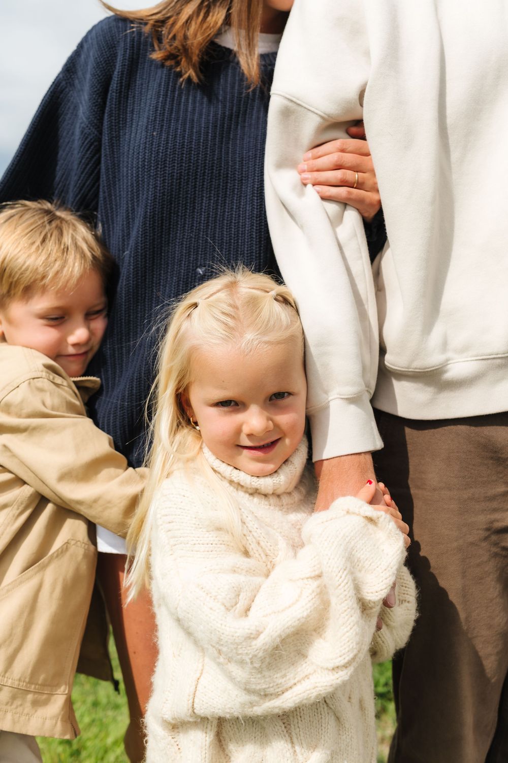 Two young blonde children smile and hug their parents outdoors in a heartwarming family portrait.