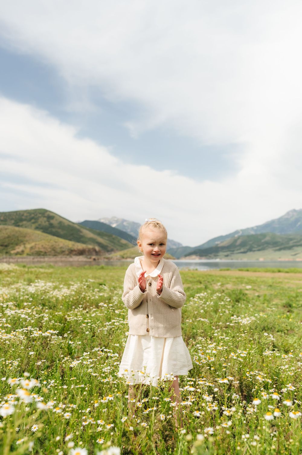 A joyful little girl in a white dress stands in a daisy field with mountains and a lake in the background.