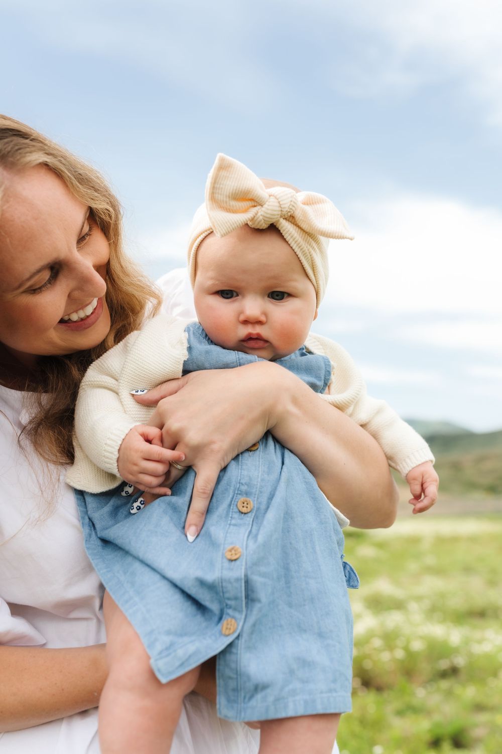 A smiling mother holds her baby girl wearing a denim dress and bow headband outdoors in a green field.