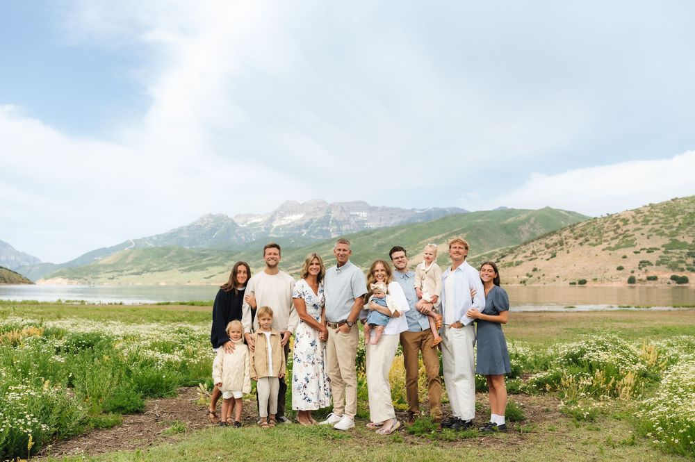 Large family posing outdoors in a green meadow with mountains and a lake in the background on a sunny day.