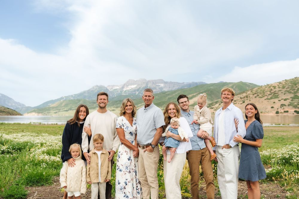 Large family group portrait outdoors with mountain lake backdrop, smiling adults and children in coordinated neutral outfits.