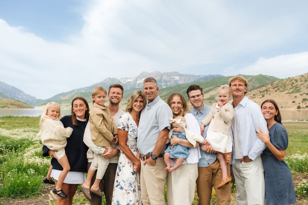 Large family group portrait outdoors with mountain backdrop, smiling adults and children posing together on a green meadow.