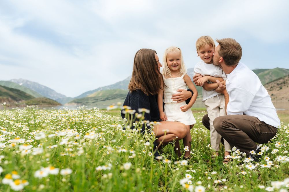 Family of four laughing together in a daisy field with mountain backdrop on a sunny day.