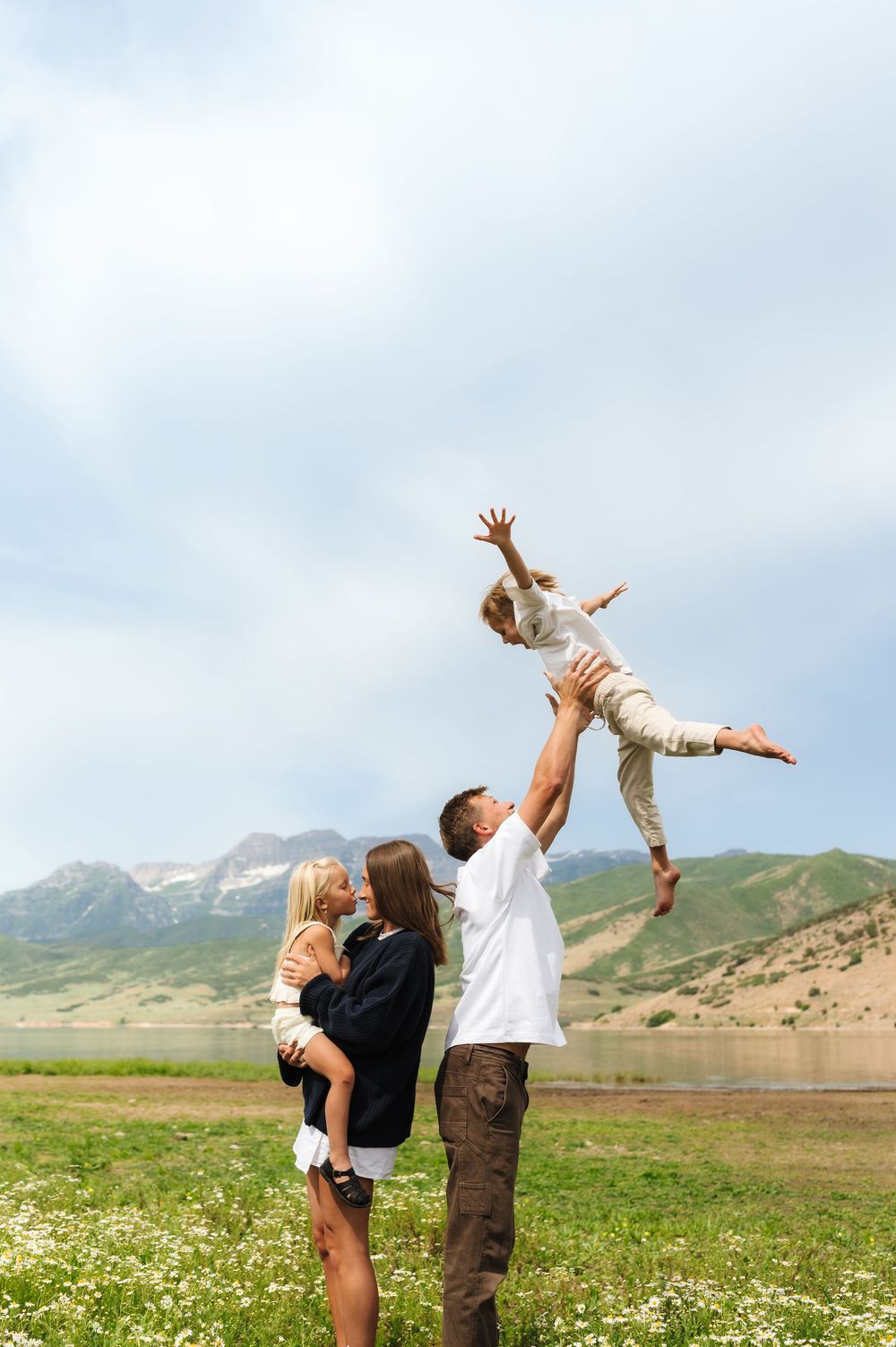 Happy family outdoors, father tossing child in air while mother holds another child, green meadow and mountains backdrop.